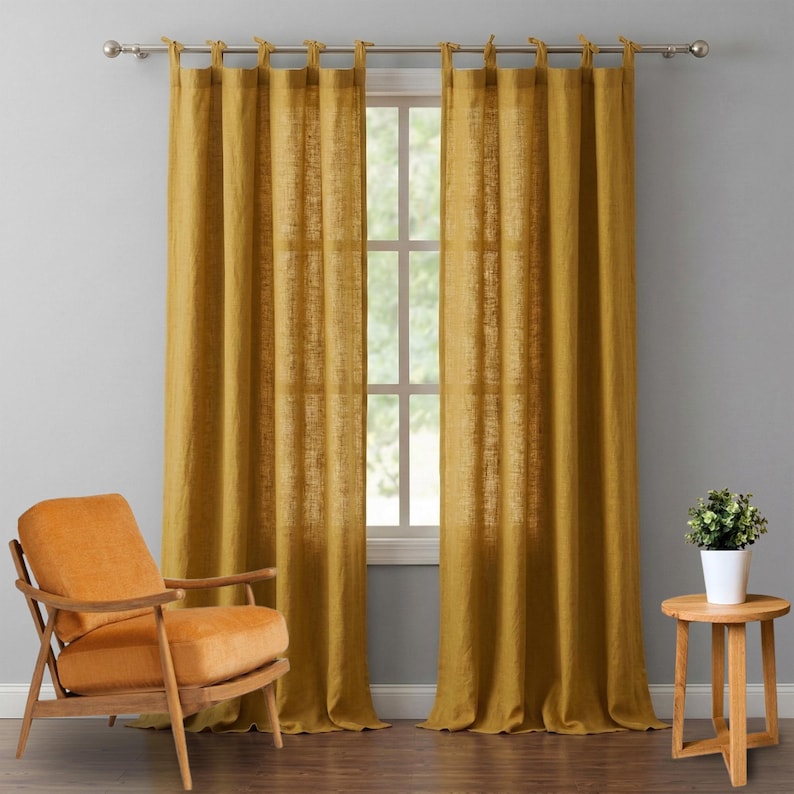 May include: Mustard yellow linen curtains hanging from a silver rod, framing a window. An orange armchair sits to the left, and a small wooden side table with a potted plant is on the right. The curtains are floor-length and have a textured appearance.