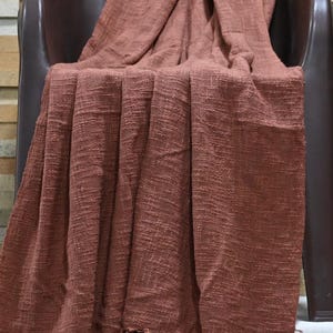 May include: A rust-colored woven throw blanket with a fringed edge is draped over a brown chair. The blanket has a textured appearance and is made of a soft material. The throw blanket is a home decor item.