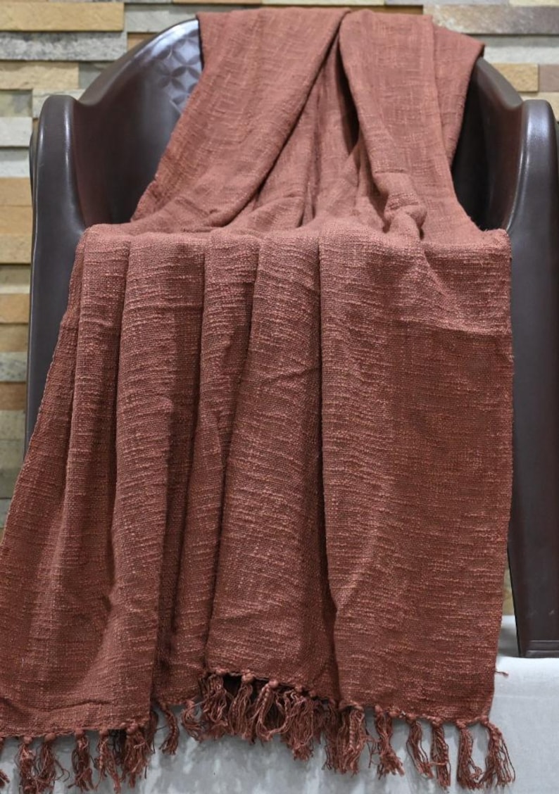 May include: A rust-colored woven throw blanket with a fringed edge is draped over a brown chair. The blanket has a textured appearance and is made of a soft material. The throw blanket is a home decor item.