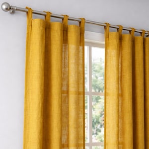 May include: Mustard yellow linen curtains hanging from a silver rod. The curtains are tab-top style, allowing them to be easily hung. The window behind the curtains is visible, with a view of trees and natural light.