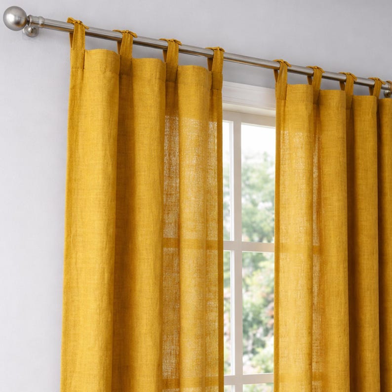May include: Mustard yellow linen curtains hanging from a silver rod. The curtains are tab-top style, allowing them to be easily hung. The window behind the curtains is visible, with a view of trees and natural light.
