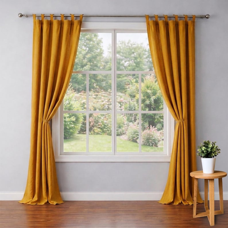 May include: Mustard yellow tab-top curtains framing a large window. The curtains are tied back, revealing a view of a lush green garden. A small wooden side table holds a potted plant, adding a touch of nature to the room.