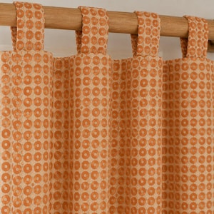 May include: A tab-top curtain panel featuring a repeating geometric pattern of orange circles on a light beige background. The fabric hangs from a wooden curtain rod, displaying vertical folds and a consistent, uniform circular print design.
