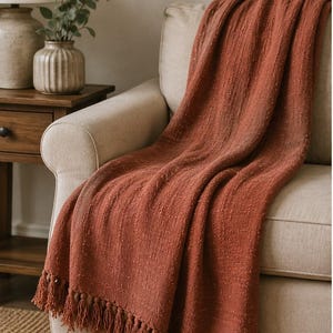May include: A terracotta-colored throw blanket with a textured weave and fringe detail draped over a beige armchair. The blanket's warm hue complements the neutral tones of the furniture, creating a cozy and inviting aesthetic. The fringe adds a decorative element.