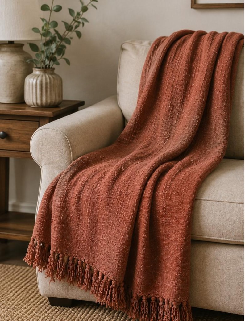 May include: A terracotta-colored throw blanket with a textured weave and fringe detail draped over a beige armchair. The blanket's warm hue complements the neutral tones of the furniture, creating a cozy and inviting aesthetic. The fringe adds a decorative element.