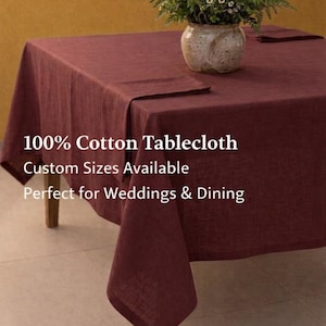 May include: A burgundy 100% cotton tablecloth draped over a wooden table. The tablecloth has a textured appearance and is paired with a vase of flowers. Text on the image reads: 100% Cotton Tablecloth, Custom Sizes Available.