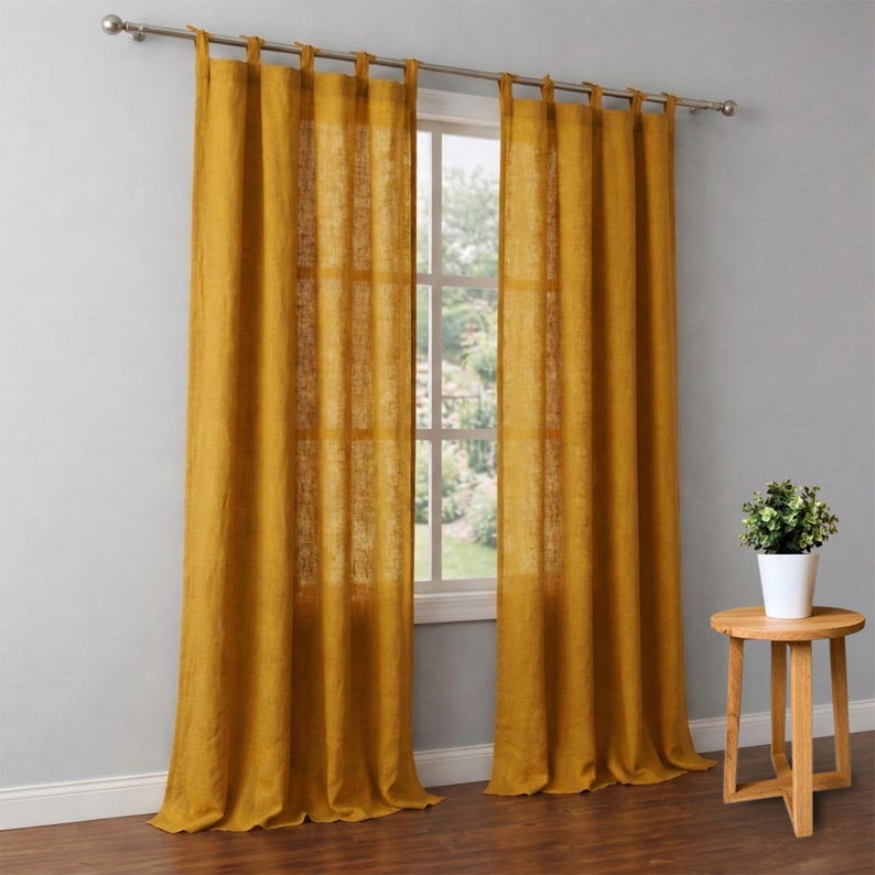 May include: Mustard yellow linen curtains hanging from a silver rod, framing a window. The curtains are long and flowing, with a slightly sheer appearance. A small wooden side table with a potted plant sits to the right of the window, adding a touch of greenery to the room.