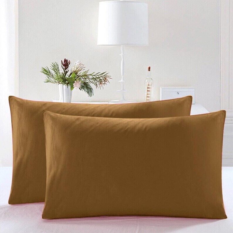 May include: Two brown pillowcases, one on top of the other, with a white lamp and a vase of flowers in the background.