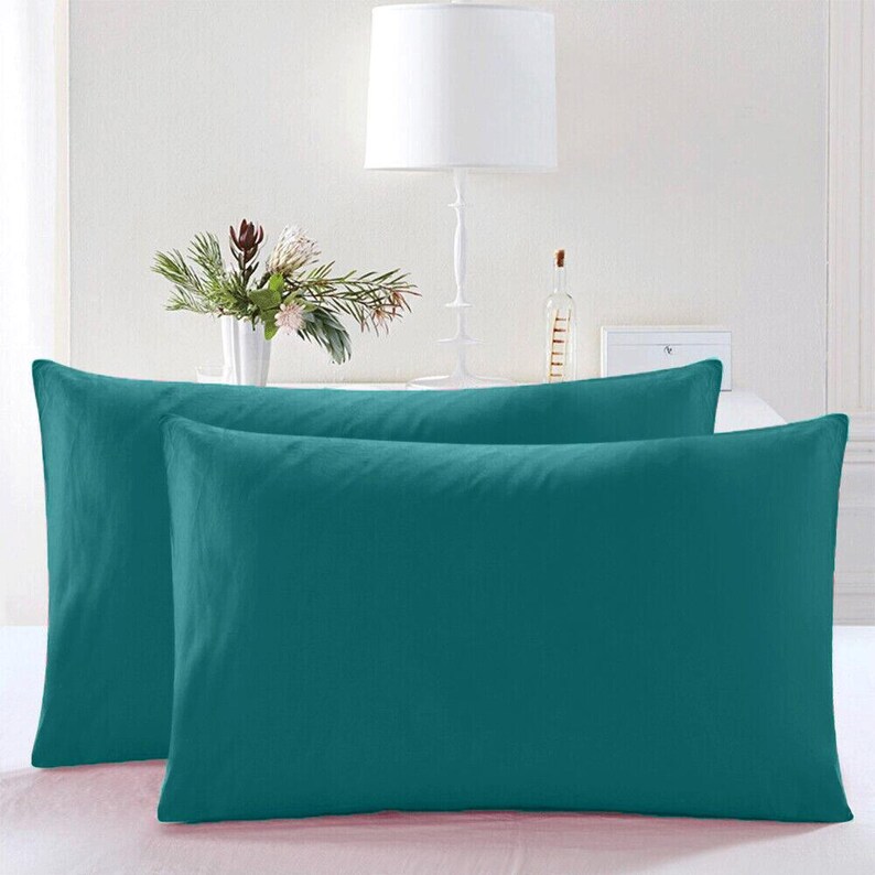 May include: Two teal blue pillowcases, one on top of the other, with a white lamp and a vase of flowers in the background.