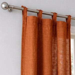 May include: Burnt orange linen curtains suspended from a brushed nickel curtain rail. The curtains feature tab-top loops for hanging. The rail has a decorative ball finial. The curtains are partially drawn, with a window visible in the background.