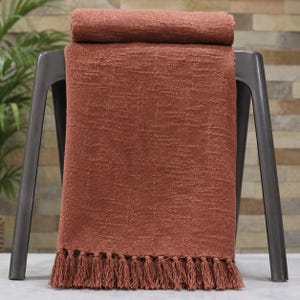 Pode incluir: Uma manta cor de terracota com uma trama texturizada e uma borda de franjas. A manta está drapeada sobre um banco de metal cinza escuro. Uma seção enrolada da manta repousa no encosto do banco.