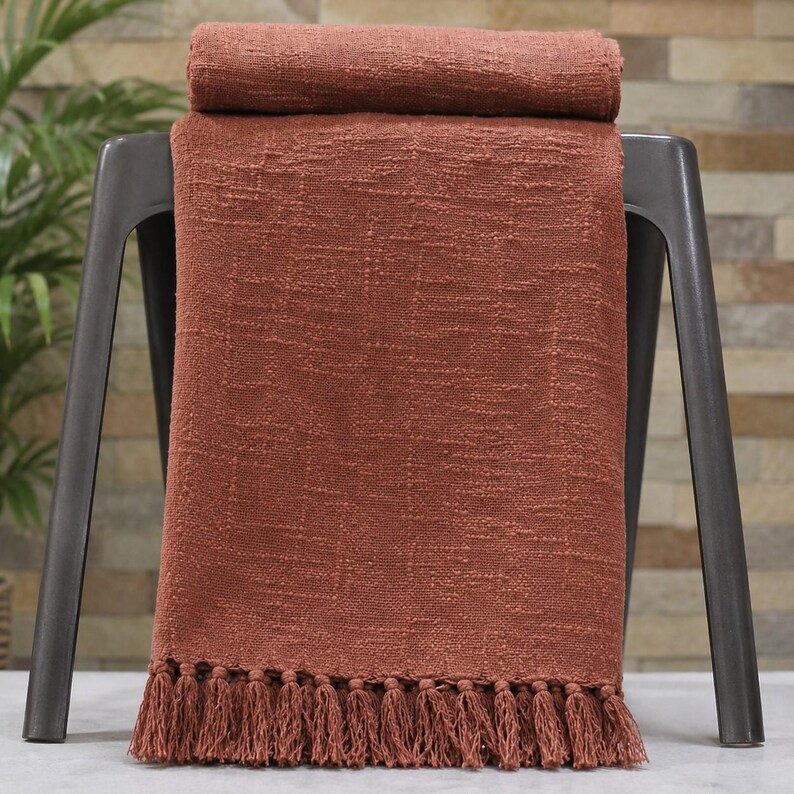 May include: A terracotta-colored throw blanket with a textured weave and a fringe trim. The blanket is draped over a dark gray metal stool. A rolled-up section of the blanket rests on the stool's back.