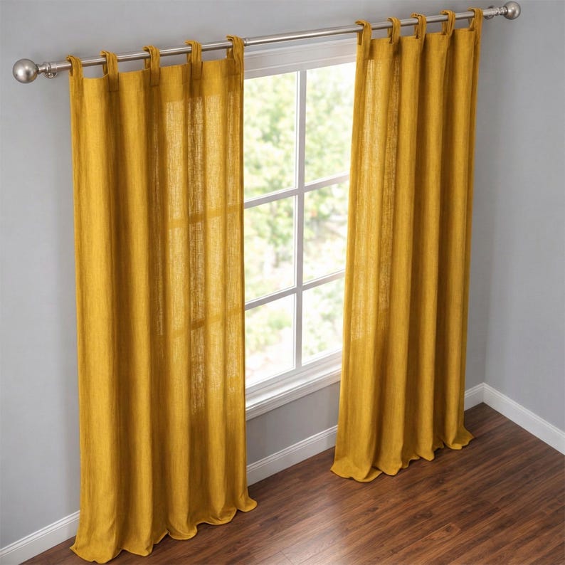 May include: Two mustard-yellow linen curtains hang from a silver rod, framing a window. The curtains are made of a textured fabric and have tab tops. The window reveals a view of green foliage outside.