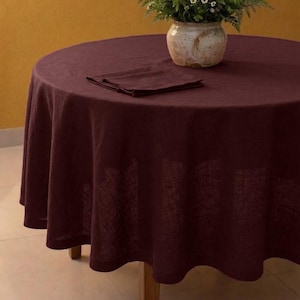 May include: A round burgundy linen tablecloth draped over a wooden table. A matching napkin is folded on the table. A ceramic vase with a bouquet of greenery and small white and purple flowers sits in the center.
