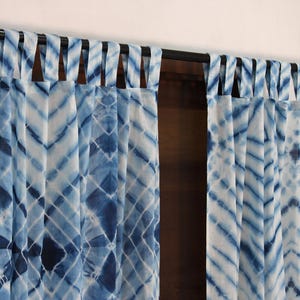 Op de afbeelding: Een paar blauwe en witte tie-dye gordijnen met een eenvoudig ontwerp. De gordijnen hangen aan een zwarte stang en hebben een geplooide look.