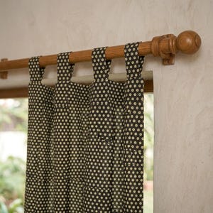 May include: A dark brown curtain with a repeating pattern of small cream-colored dots, featuring fabric tabs at the top that hang from a smooth, light-toned wooden curtain rod with rounded finials mounted on a textured off-white wall.