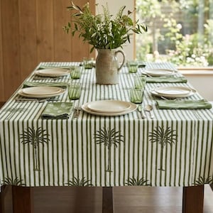 May include: A dining table set for a meal. The table is covered with a white and green striped tablecloth with palm tree designs. Beige plates, green glasses, and napkins are set for each place setting. A vase of greenery sits in the center.