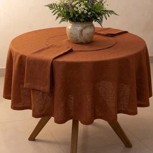 May include: A circular, terracotta-coloured linen tablecloth with a matching napkin and placemat. A ceramic vase with a floral arrangement sits on the table. The table has a wooden base.