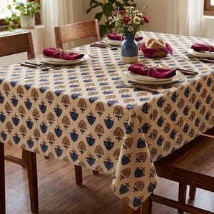 May include: A patterned tablecloth in beige, blue, and brown covers a wooden dining table. The table is set with plates, napkins, and cutlery. A blue vase holds flowers, and a basket of bread is placed on the table. The table is surrounded by wooden chairs.