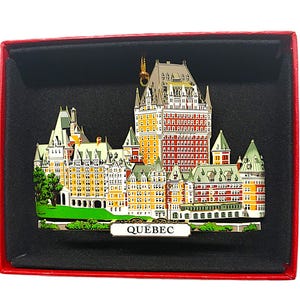 May include: A red and gold metal ornament depicting the Chateau Frontenac in Quebec City. The ornament is in a red box with a black interior and the word "QUEBEC" printed on the front.