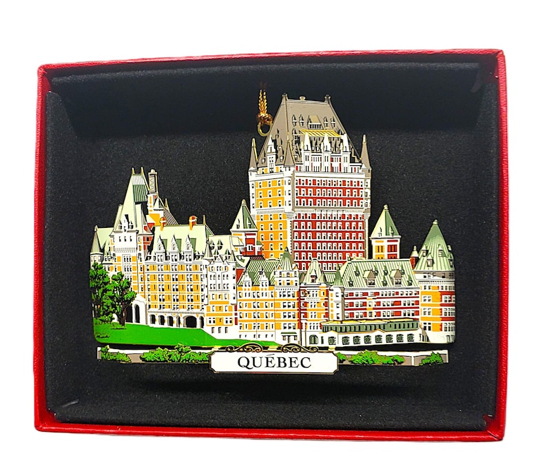 May include: A red and gold metal ornament depicting the Chateau Frontenac in Quebec City. The ornament is in a red box with a black interior and the word "QUEBEC" printed on the front.