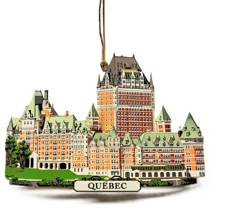 May include: A wooden ornament depicting the Chateau Frontenac hotel in Quebec City, Canada. The ornament is painted in shades of brown, green, and white, with the words "QUEBEC" printed on a banner below the building.