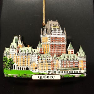 May include: A gold-colored metal ornament depicting the Chateau Frontenac hotel in Quebec City. The ornament is detailed with red, white, and green paint and features the word "QUEBEC" on a sign at the bottom.