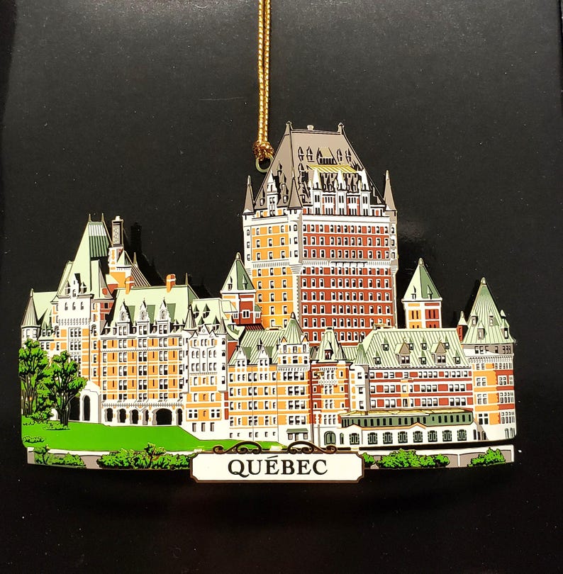 May include: A gold-colored metal ornament depicting the Chateau Frontenac hotel in Quebec City. The ornament is detailed with red, white, and green paint and features the word "QUEBEC" on a sign at the bottom.
