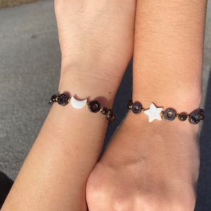 May include: Two bracelets with black beads and gold accents. One bracelet features a white crescent moon charm, and the other features a white star charm.