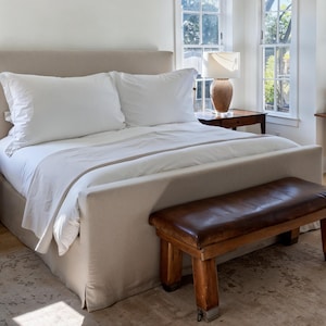 Könnte beinhalten: Ein beige gepolstertes Bett mit weißer Bettwäsche und Kissen. Am Fußende des Bettes befindet sich eine braune Lederbank. Ein Nachttisch aus Holz mit einer Lampe und ein Stuhl befinden sich ebenfalls im Raum. Der Raum hat große Fenster.