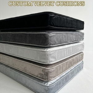 May include: A stack of custom velvet cushions in various shades of gray, from dark charcoal to light silver. The cushions have a rectangular shape with a piped edge and are made of a soft, plush fabric. The text "CUSTOM VELVET CUSHIONS" is displayed at the top.