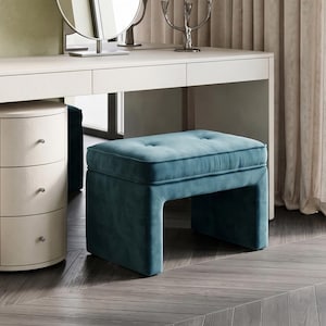 Op de afbeelding: Een rechthoekige, blauwgroene fluwelen poef met een gecapitonneerde zitting met twee knopen en een solide, doorlopend onderstel. De bekleding heeft een zachte, matte textuur en het meubelstuk staat voor een witte kaptafel met lades.