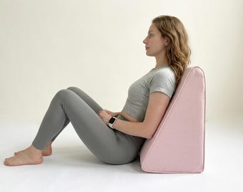 May include: A light pink wedge-shaped pillow with a textured fabric cover. The pillow features a triangular profile designed to provide back support while sitting in an upright position on the floor, maintaining a firm and structured shape.