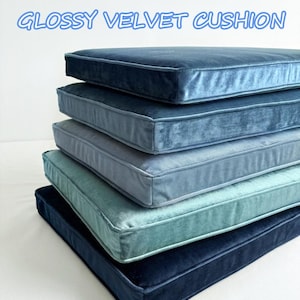 May include: A stack of five square velvet cushions in shades of blue, from light teal to deep navy. The cushions have a glossy sheen and a simple, tailored design. The text "GLOSSY VELVET CUSHION" is displayed in blue above the cushions.