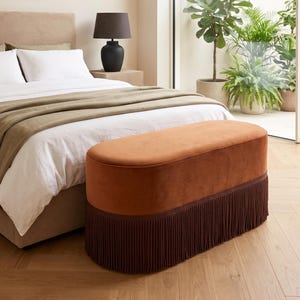 May include: An oval-shaped bench upholstered in burnt orange velvet fabric, featuring a dark brown fringe trim along the base. The bench has a smooth, rounded top surface and a structured, elongated form that sits low to the ground.
