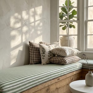 May include: A window seat with a green and white striped cushion and several decorative pillows. The pillows feature patterns like gingham and botanical prints. The wooden frame of the seat is visible, and a small white table and vase are in the foreground.