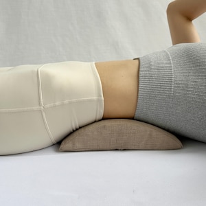 May include: A person lying on their back with a brown lumbar support pillow under their lower back. They are wearing white leggings and a grey ribbed tank top.