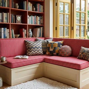 Könnte beinhalten: Eine gemütliche Leseecke mit einer Eckbank mit rot-weiß gestreiften Kissen und dekorativen Kissen. Im Hintergrund ein Bücherregal aus Holz mit Büchern und eine kleine Uhr. Ein flauschiger weißer Teppich und gelb gerahmte Türen vervollständigen den einladenden Raum.