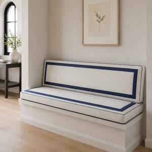 May include: A white built-in bench seat featuring a long rectangular backrest and a matching seat cushion. Both cushions are upholstered in an off-white fabric and are accented with a bold, dark navy blue rectangular border trim design.