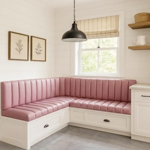 May include: An L-shaped corner banquette seating unit featuring dusty pink vertical channel-tufted upholstery atop a white wooden base with integrated storage drawers, accented by dark metal hardware pulls in a bright, white-walled room.