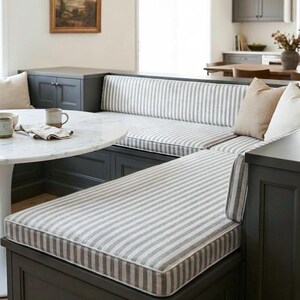 May include: A built-in dining bench with gray and white striped cushions. The bench is dark gray and surrounds a white marble table. A framed landscape painting hangs on the wall above the bench.