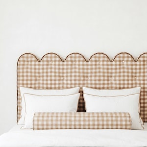 May include: A scalloped headboard upholstered in a tan and white gingham check fabric with tufted buttons and a brown piped edge. The bed features two white rectangular pillows with tan piping and a matching tan and white gingham bolster pillow.