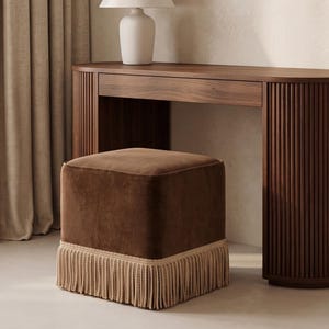 May include: A square ottoman upholstered in dark brown velvet fabric featuring a thick beige rope fringe trim along the bottom edge. The ottoman is positioned in front of a dark wood vanity table with a fluted design and a white table lamp.