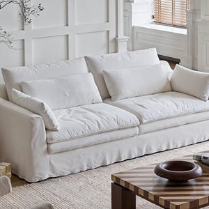 May include: A white two-seater sofa with several white cushions. The sofa has a skirted base and is in a room with white paneling and a light-coloured rug. A wooden coffee table and a floor lamp are also visible.