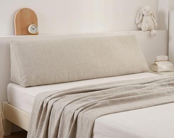 May include: A large, wedge-shaped bed pillow upholstered in a textured, light beige fabric. The pillow features a slanted back design and sits horizontally across the width of a bed, which is covered with a matching light beige linen-style blanket.