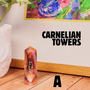May include: A carnelian tower-shaped crystal with orange, red, and white hues. The crystal has a polished surface and a unique internal pattern. The text "CARNELIAN TOWERS" and the letter "A" are visible in the image.