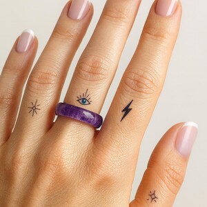 May include: A hand with French-manicured nails and small tattoos of a starburst, an eye, and a lightning bolt. A purple ring is worn on the ring finger. The skin tone is light.