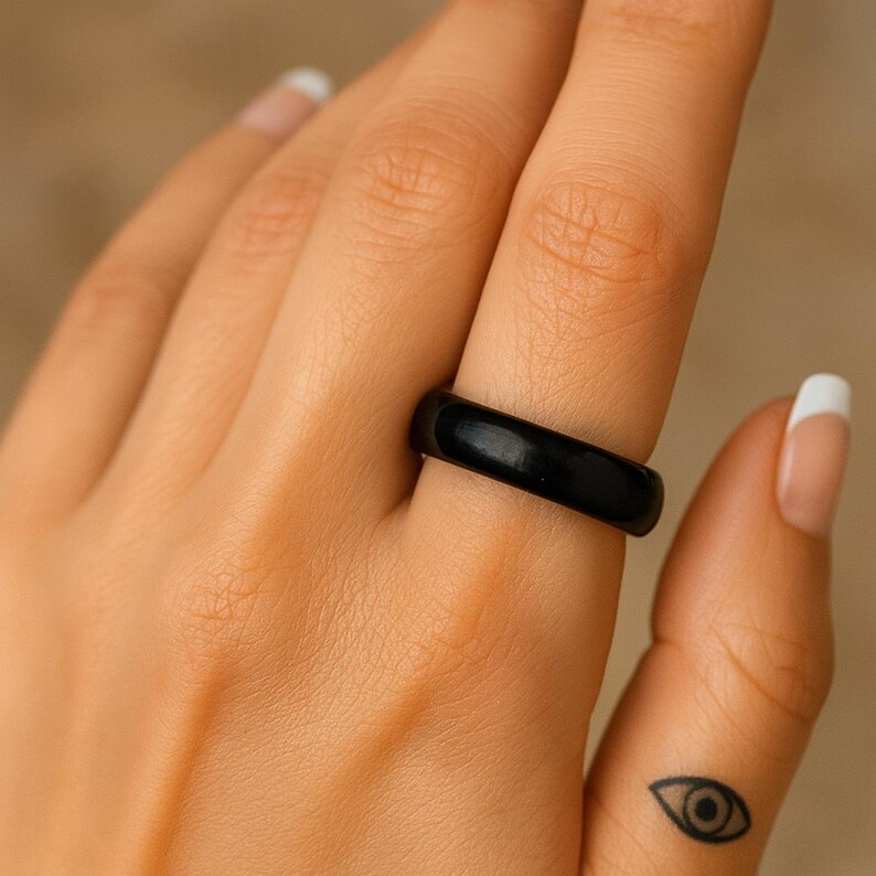 May include: A black ring is worn on a finger. The ring is a solid, glossy black color. The ring is a simple band design. The person's hand has a small eye tattoo on the finger.
