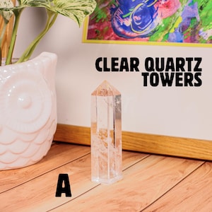 May include: A clear quartz tower stands on a wooden surface. The tower is rectangular with a pointed top. The text "CLEAR QUARTZ TOWERS" is displayed in black. A pink owl-shaped planter and a framed artwork are in the background.
