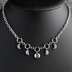 May include: A silver-toned necklace with a chain and crescent moon charms, along with a central circular pendant. The necklace is displayed on a black mannequin, set against a textured gray background.